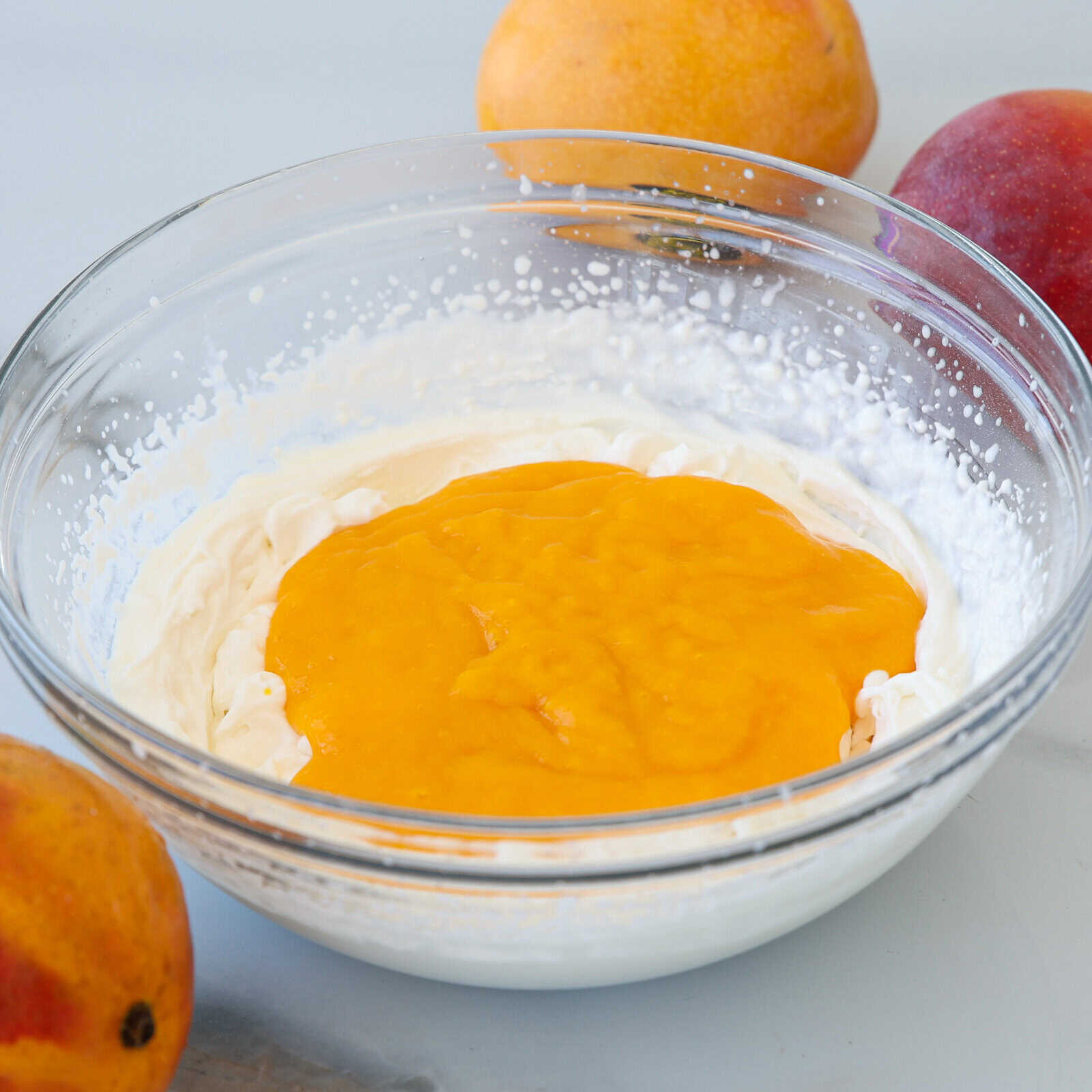 A glass bowl filled with whipped cream and a layer of bright orange mango puree, surrounded by whole ripe mangoes on a light surface.