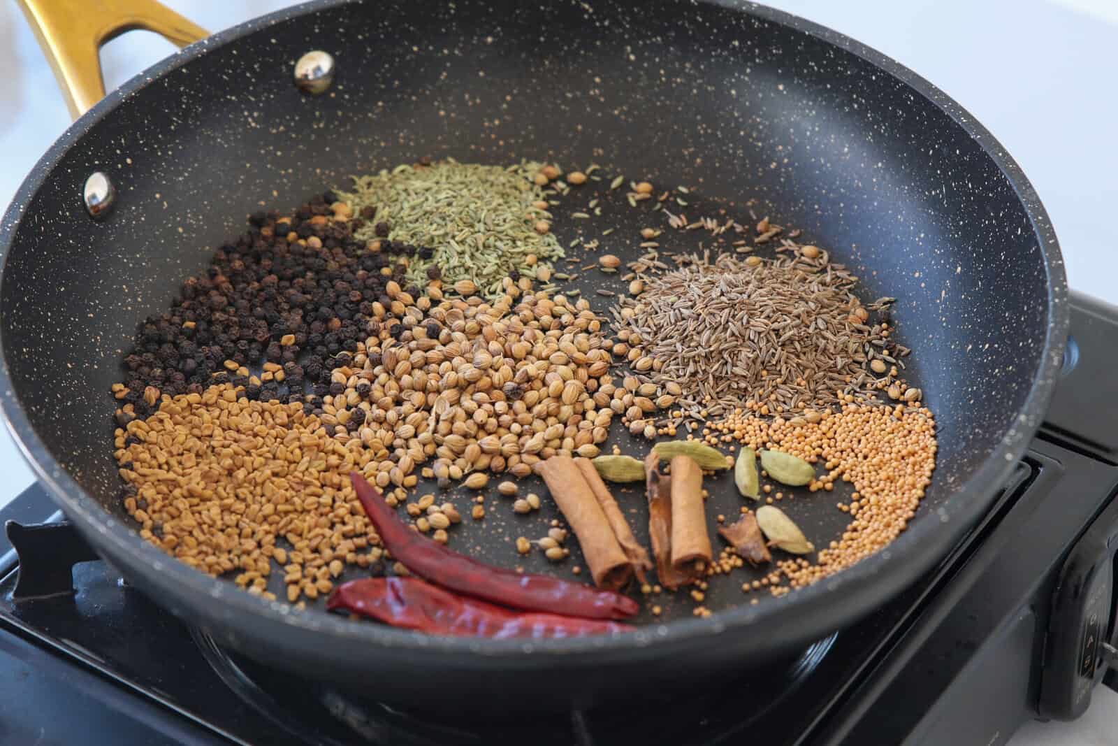 A nonstick pan on a stove contains whole spices, including dried red chilies, cinnamon sticks, cardamom pods, black peppercorns, coriander seeds, cumin seeds, fennel seeds, mustard seeds, and fenugreek seeds.