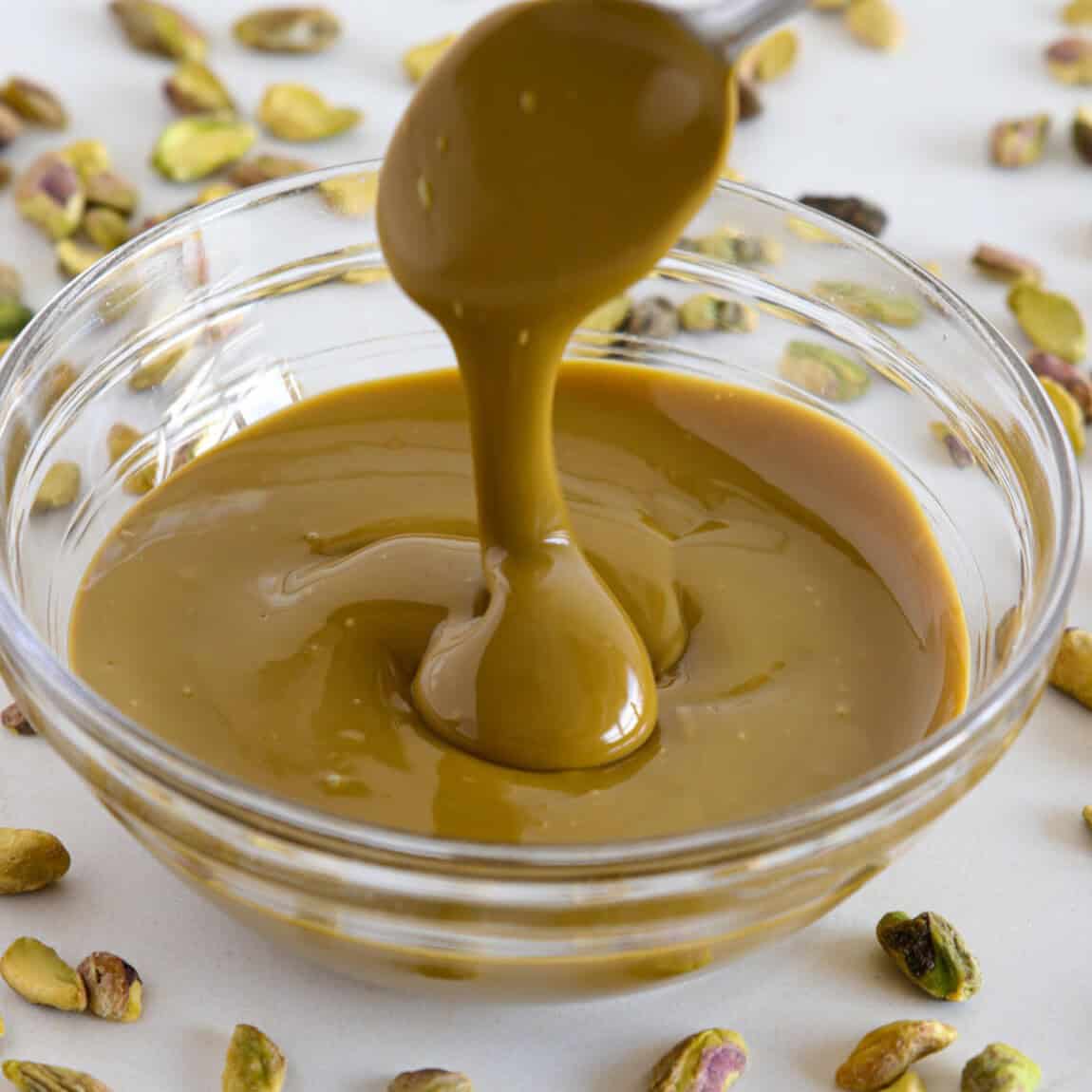 A glass bowl filled with creamy pistachio paste, with a spoon lifting and letting the thick paste drizzle down, surrounded by scattered whole pistachios on a white surface.