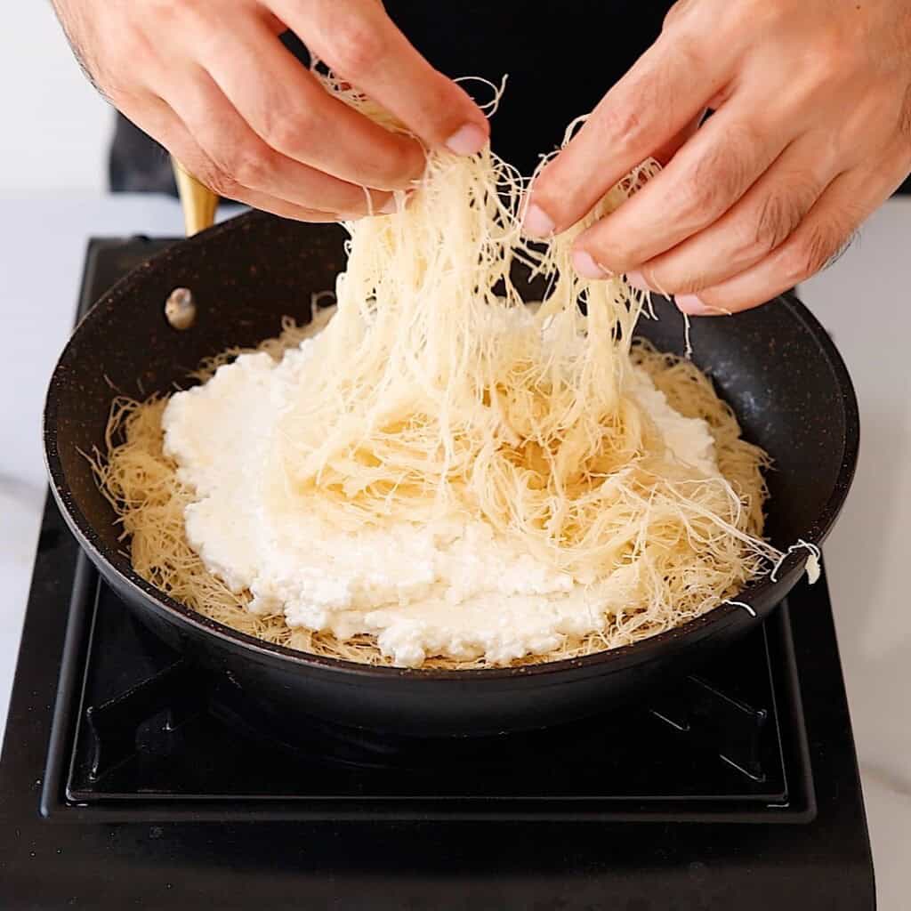 A person’s hands layering shredded pastry dough over a pan filled with a creamy white mixture, preparing a dish on a stovetop.