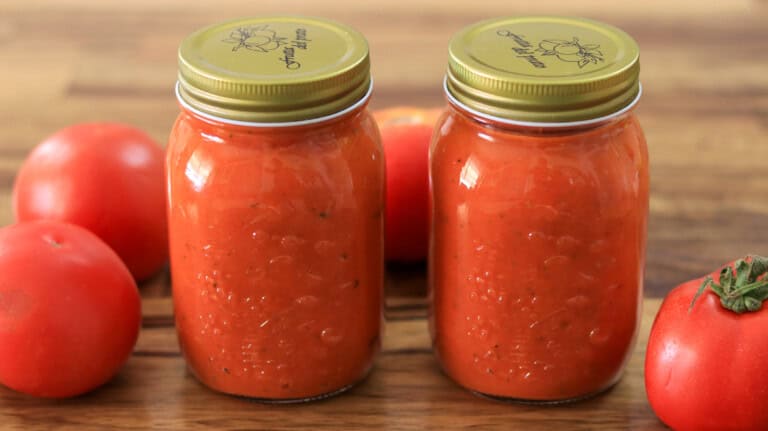Two glass jars filled with homemade tomato sauce, sealed with gold lids, are placed on a wooden surface. Fresh red tomatoes are arranged around the jars.