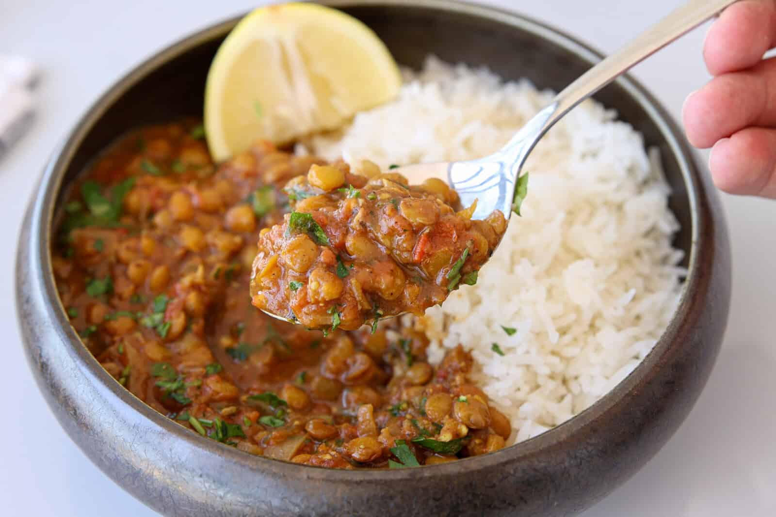A bowl of white rice and lentil curry garnished with herbs, served with a lemon wedge. A hand holds a spoonful of the lentil curry above the bowl.