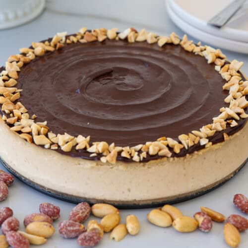A round peanut butter cheesecake topped with a smooth layer of chocolate and a ring of chopped peanuts, surrounded by scattered peanuts on a white surface. Plates and utensils are in the background.