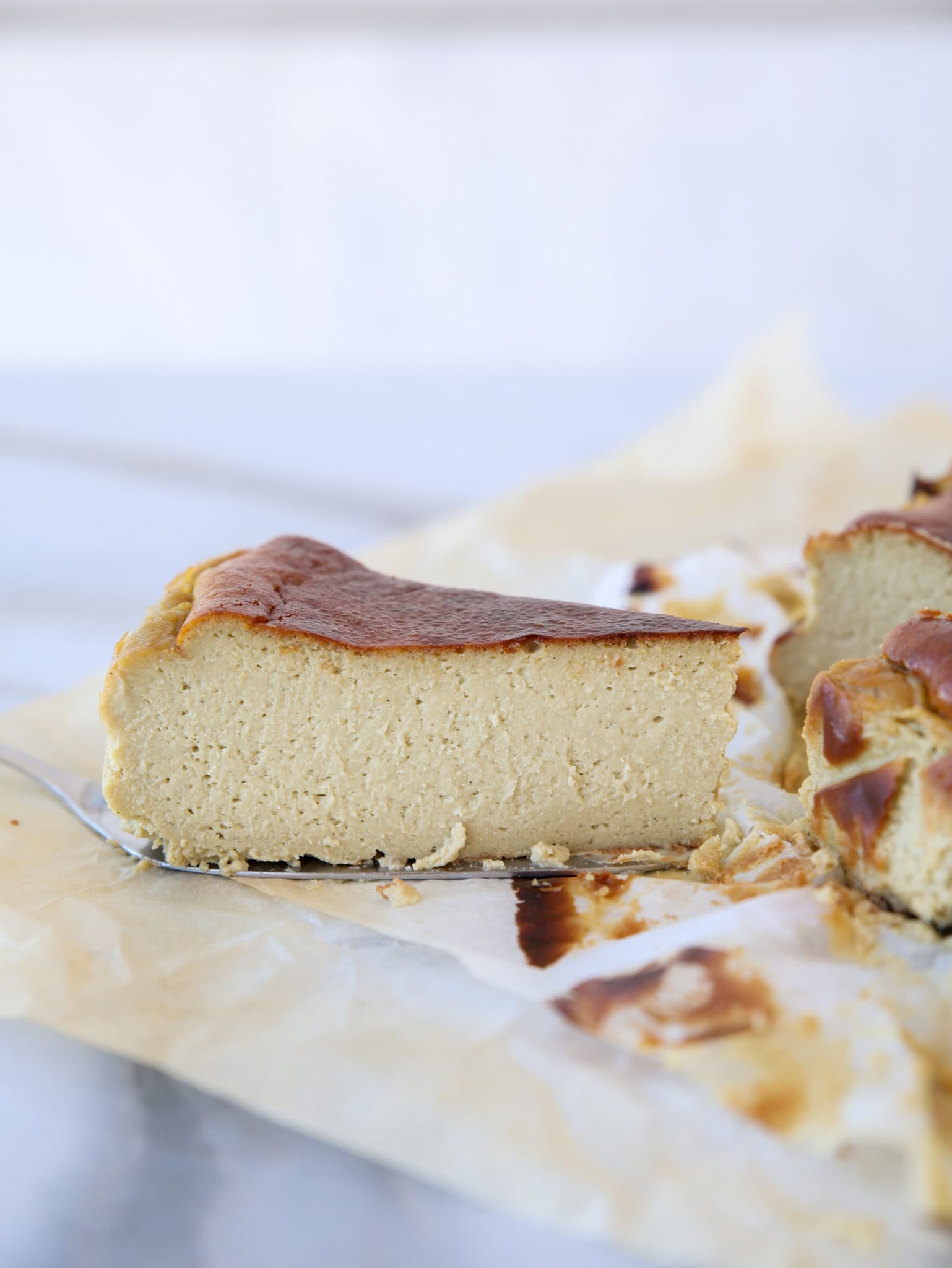 A close-up of a thick slice of cheesecake with a golden-brown top, sitting on parchment paper with the rest of the cheesecake visible in the background.