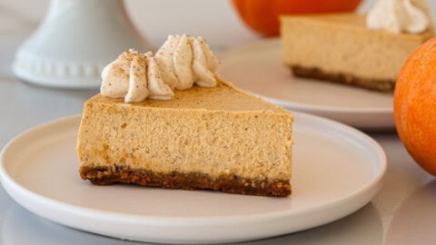A slice of a creamy pumpkin cheesecake with a graham cracker crust, topped with whipped cream and a sprinkle of spice, sits on a white plate. Another cheesecake slice and pumpkins are blurred in the background.