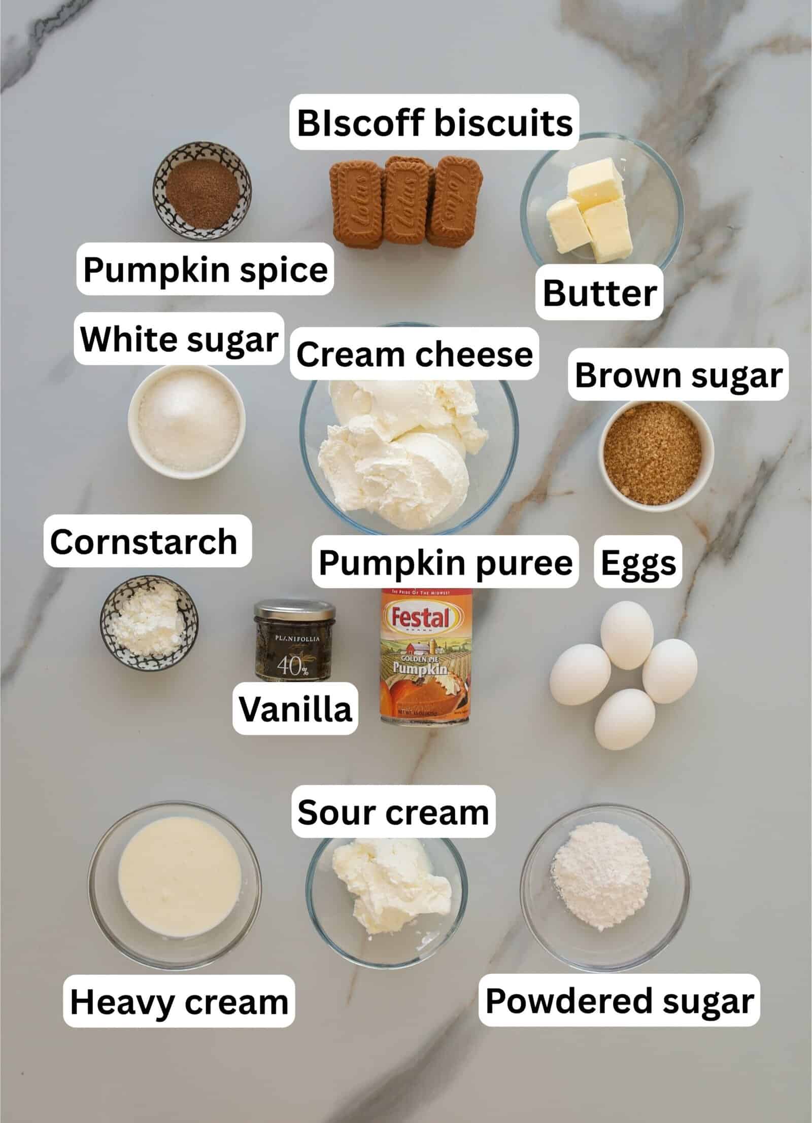 Top-down view of labeled baking ingredients on a marble surface, including Biscoff biscuits, butter, cream cheese, sour cream, heavy cream, eggs, pumpkin puree, pumpkin spice, sugars, cornstarch, vanilla, and powdered sugar.