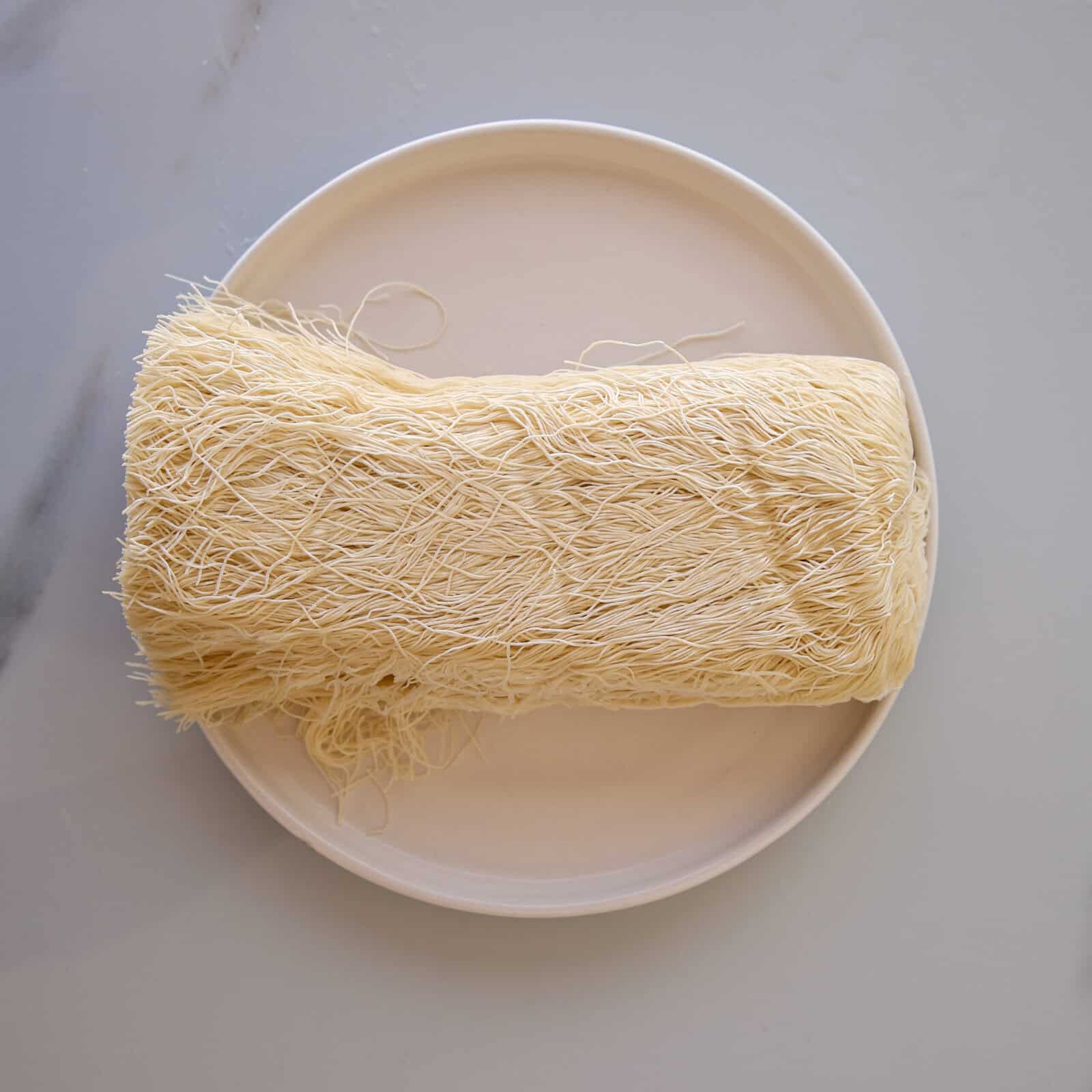 A bundle of uncooked kataifi dough, with thin, hair-like strands, sits on a round white plate against a light-colored background.