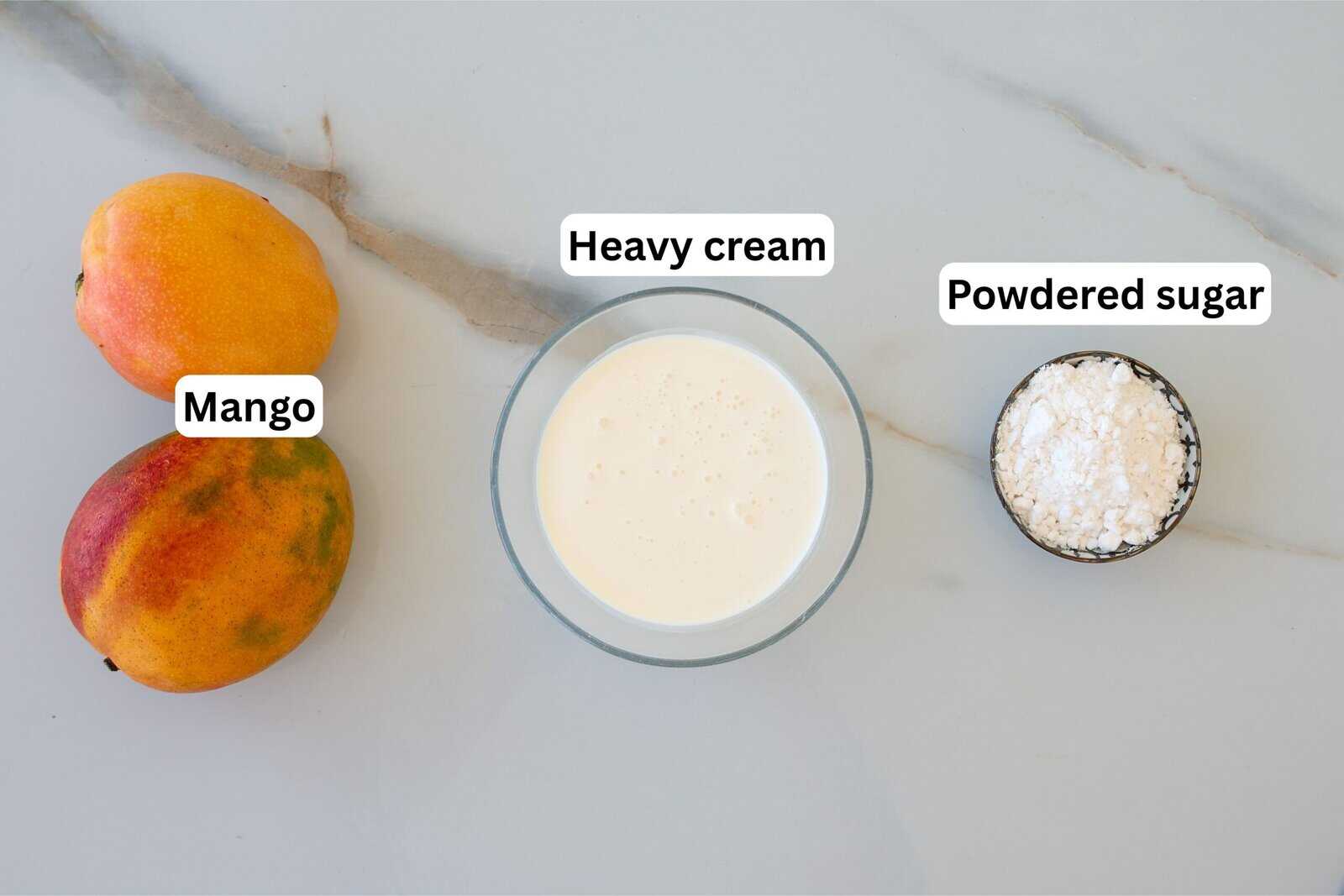 Two mangoes, a bowl of heavy cream, and a small bowl of powdered sugar are arranged on a white marble surface, each labeled with text.