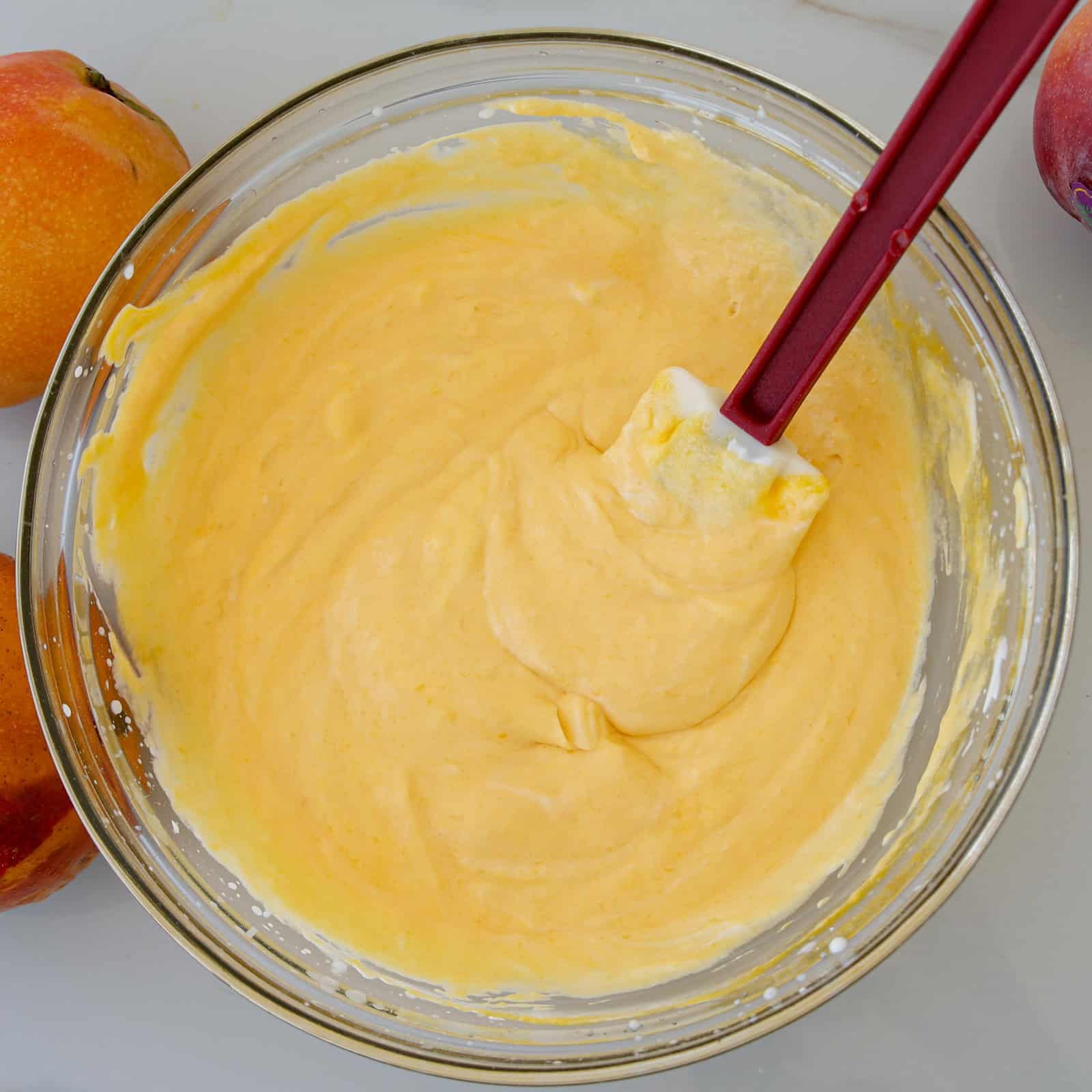 A glass bowl filled with creamy, yellow batter being stirred by a red spatula, with two whole peaches placed nearby on a white surface.