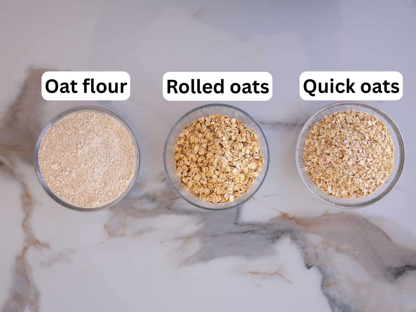 Three glass bowls on a marble surface, each containing a different oat product. From left to right: oat flour (finely ground), rolled oats (flat flakes), and quick oats (smaller flakes). Labels identify each type.