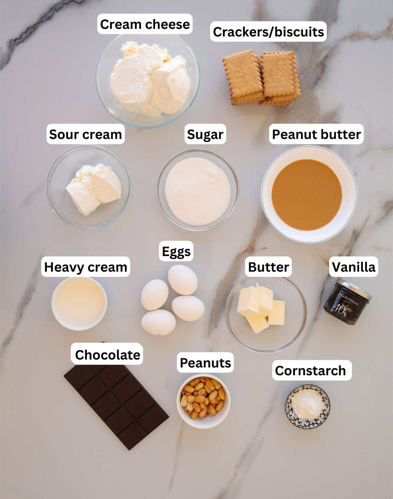Top-down view of labeled ingredients for a dessert, including cream cheese, crackers/biscuits, sour cream, sugar, peanut butter, eggs, butter, heavy cream, vanilla, chocolate, peanuts, and cornstarch on a marble surface.