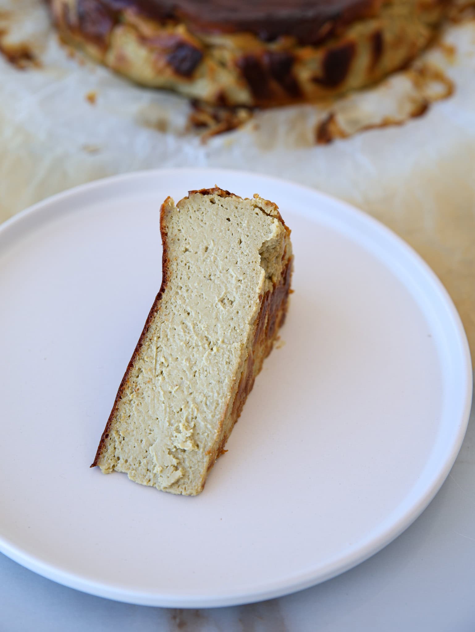 A single slice of burnt Basque cheesecake sits on a plain white plate, showing its creamy interior and browned edges. The rest of the cheesecake is blurred in the background.