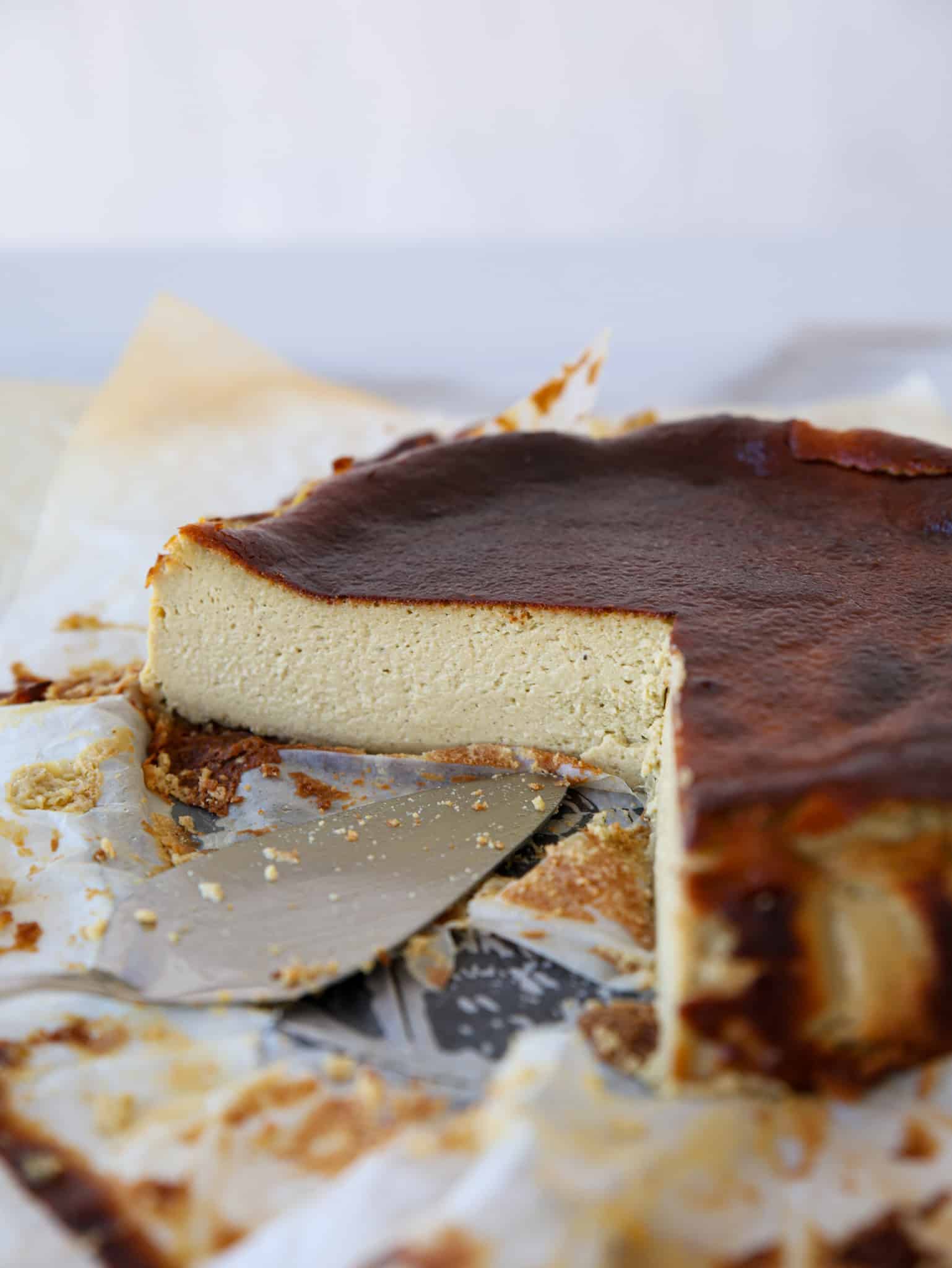 A sliced pistachio Basque burnt cheesecake sits on parchment paper, with a cake server beneath a piece. The top is dark brown and caramelized, while the inside appears creamy and smooth. Crumbs are scattered around.