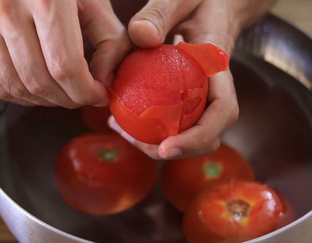 A person peels the skin off a blanched tomato over a bowl of water containing more tomatoes with loosened skins.