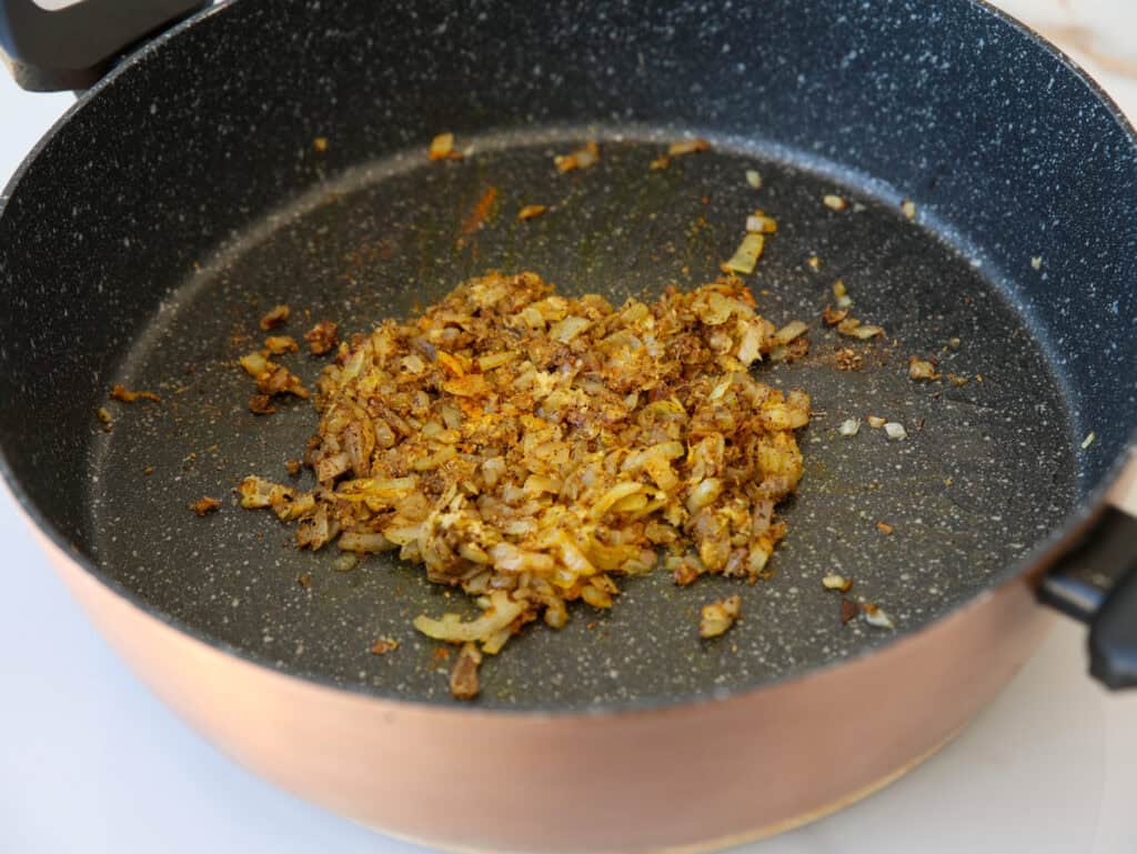 Chopped onions and spices sautéing in a large nonstick skillet, beginning to turn golden brown. The mixture is centered in the pan, with bits scattered around.