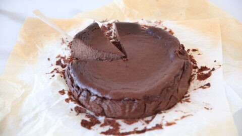 A rich, dark chocolate cake sits on parchment paper, with one slice partially lifted out by a cake server. The cake appears dense and fudgy, with crumbs scattered around.