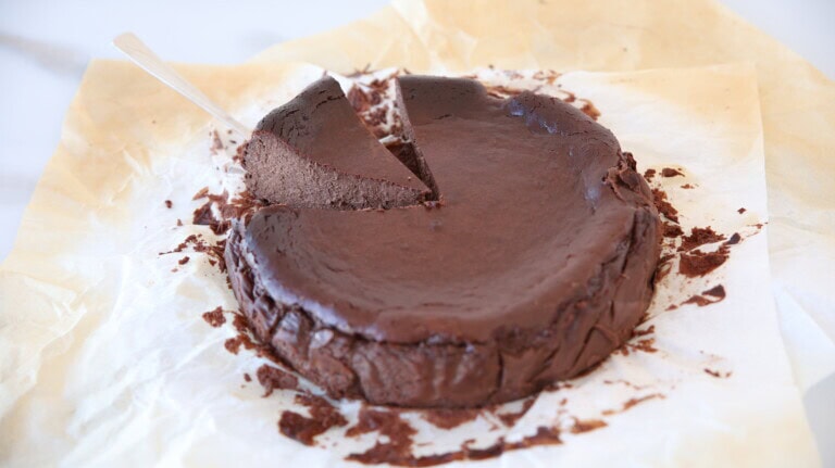 A rich, dark chocolate cake sits on parchment paper, with one slice partially lifted out by a cake server. The cake appears dense and fudgy, with crumbs scattered around.