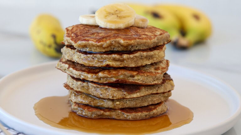 A stack of oatmeal and banana pancakes topped with banana slices sits on a white plate, with syrup drizzled around the base. In the background, whole bananas are slightly out of focus.