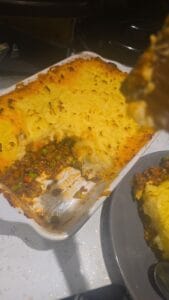 A freshly baked shepherd's pie in a white dish with a portion removed, revealing a layer of mashed potatoes on top and a filling of peas, carrots, and ground meat below. A serving is on a nearby gray plate.