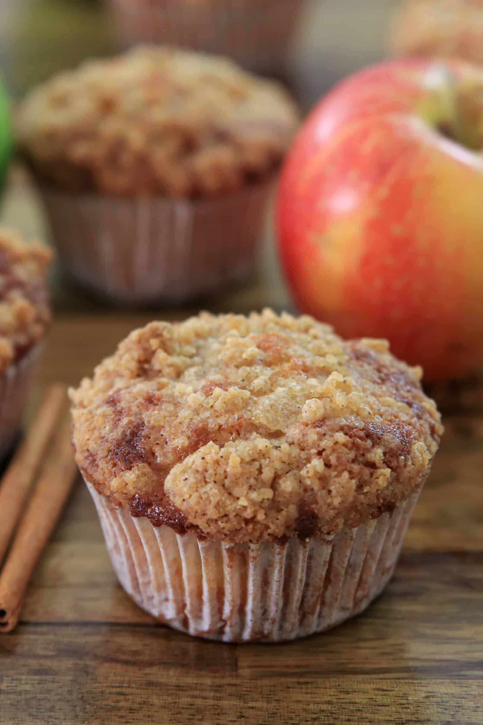 A close-up of a cinnamon-streusel muffin on a wooden surface, with cinnamon sticks and a red apple beside it. More muffins and another apple are blurred in the background.