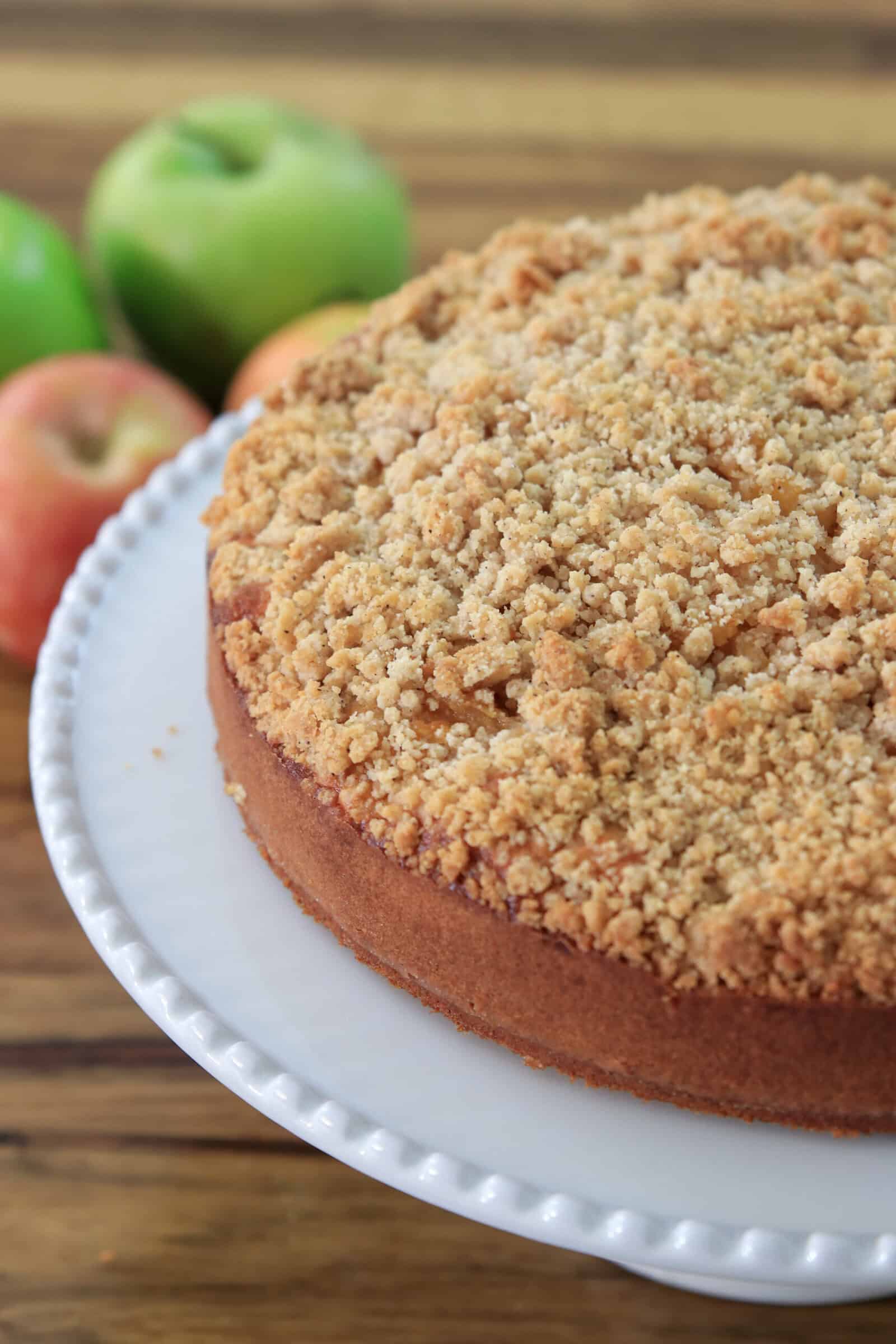 A round apple crumble cake with a golden, crumbly topping sits on a white cake stand. Green and red apples are blurred in the background on a wooden surface.