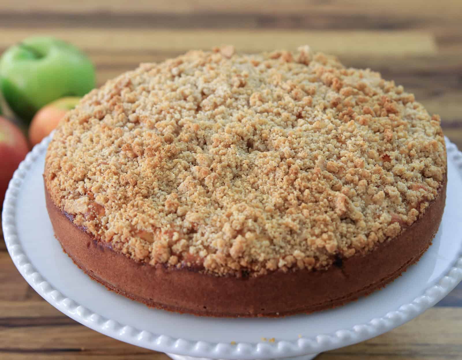 A round apple crumble cake with a golden, crumbly topping sits on a white, scalloped-edge cake stand. Fresh green apples are blurred in the background on a wooden surface.