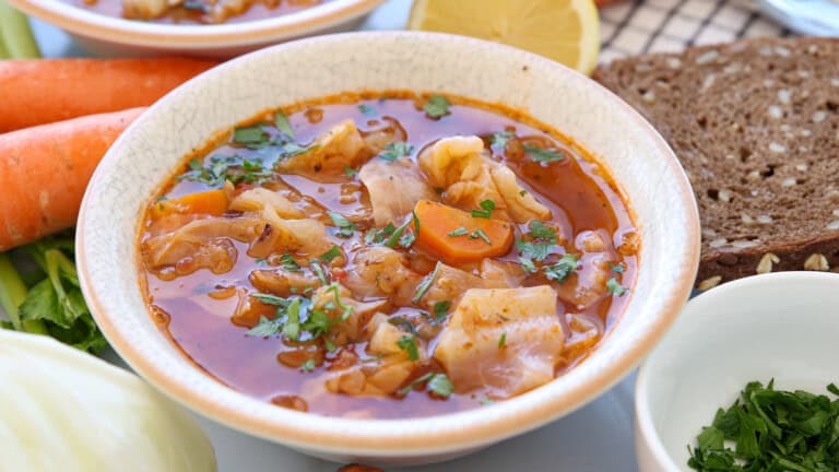 A bowl of vegetable soup with cabbage, carrots, and herbs in a tomato broth, garnished with parsley. Slices of whole grain bread, fresh carrots, cabbage, and a lemon are in the background.