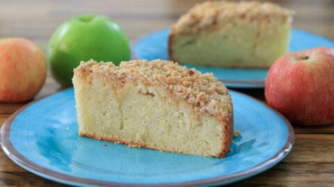 Easy Apple Crumble Cake with Streusel Topping A slice of crumbly apple cake sits on a blue plate, with green and red apples and another slice of cake visible in the background on a wooden table.