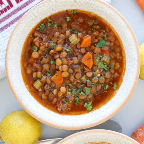 A bowl of lentil soup with chopped carrots, celery, and herbs, placed on a white surface next to a lemon, carrot, and a red-striped kitchen towel.