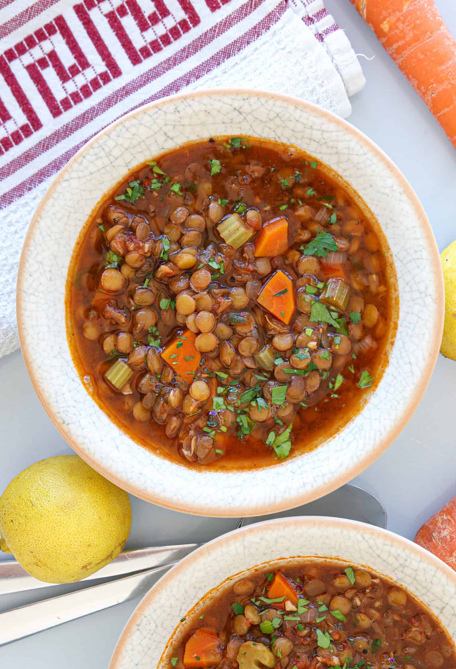 A bowl of lentil soup with chopped carrots, celery, and herbs, placed on a white surface next to a lemon, carrot, and a red-striped kitchen towel.