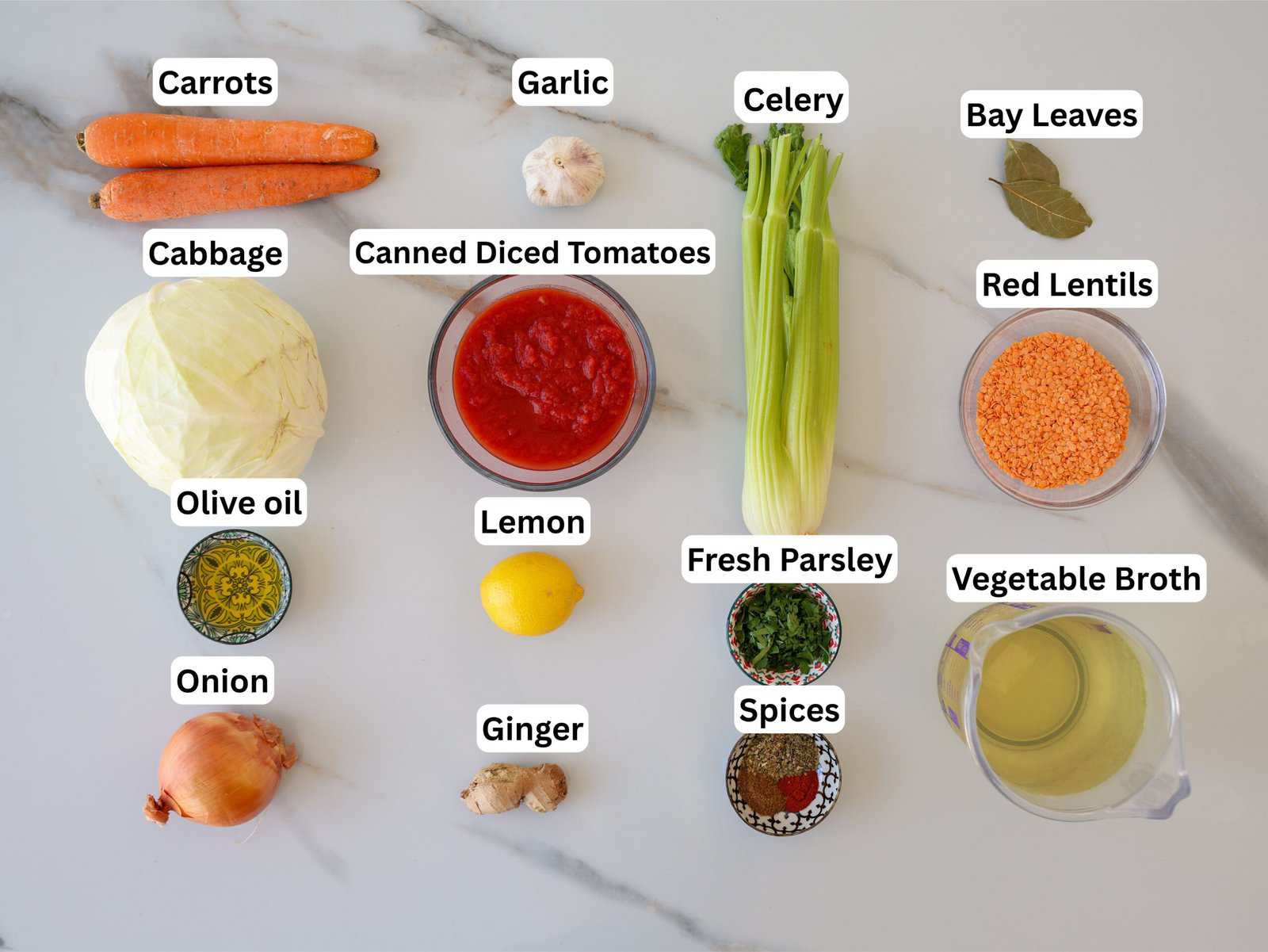 Overhead view of labeled ingredients for soup on a white surface, including carrots, garlic, celery, bay leaves, red lentils, cabbage, canned diced tomatoes, lemon, olive oil, onion, ginger, fresh parsley, spices, and vegetable broth.