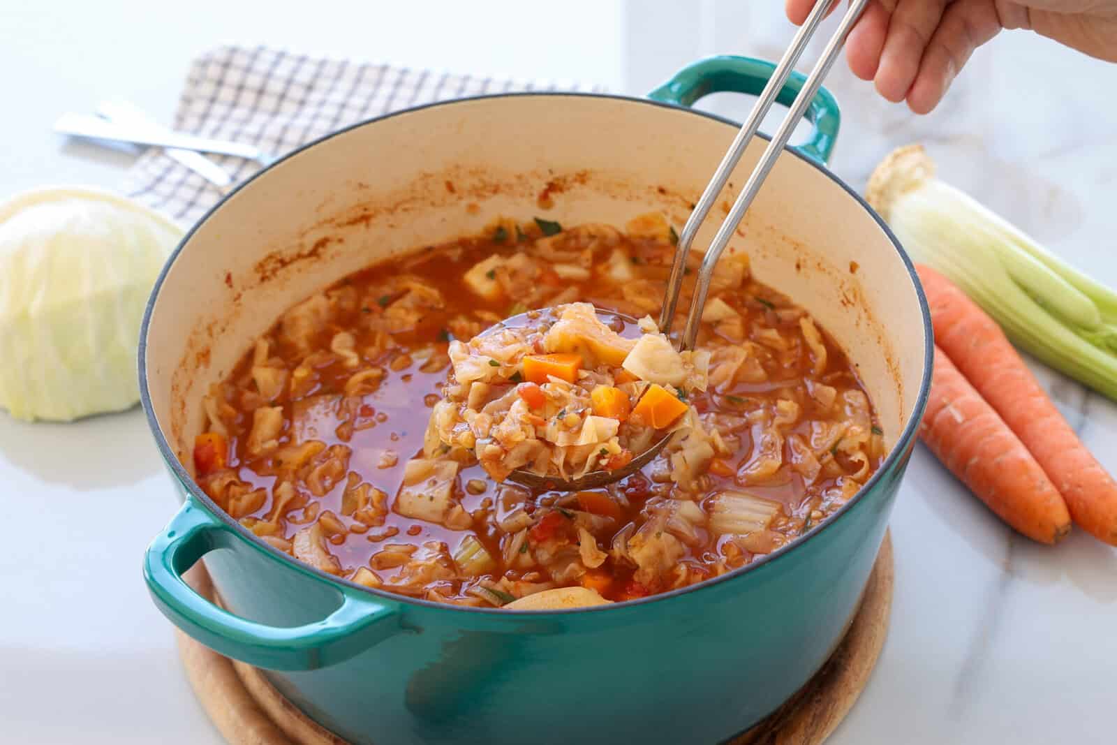 A hand holds a ladle full of vegetable soup over a teal pot. The soup contains chunks of cabbage, carrots, potatoes, and tomatoes. Fresh vegetables and a checked cloth are visible in the background.