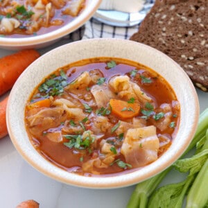 A bowl of cabbage soup with carrots and herbs, served next to slices of dark bread, fresh carrot, and celery on a white table.