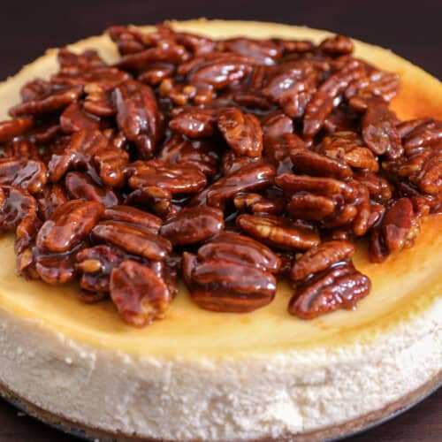A cheesecake topped with a glossy layer of caramelized pecans, sitting on a dark surface.