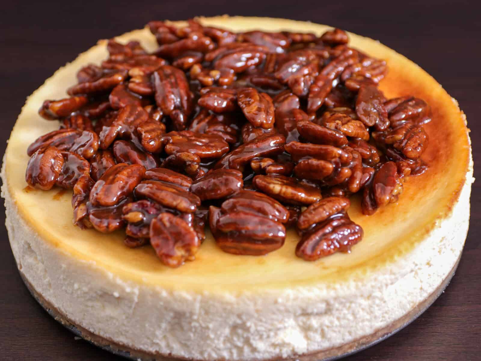 A cheesecake topped with a glossy layer of caramelized pecans, sitting on a dark surface.