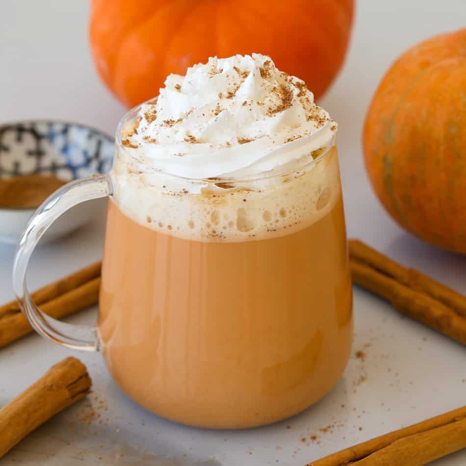 A glass mug of pumpkin spice latte topped with whipped cream and cinnamon, surrounded by cinnamon sticks, pumpkins, and a small bowl of ground cinnamon.