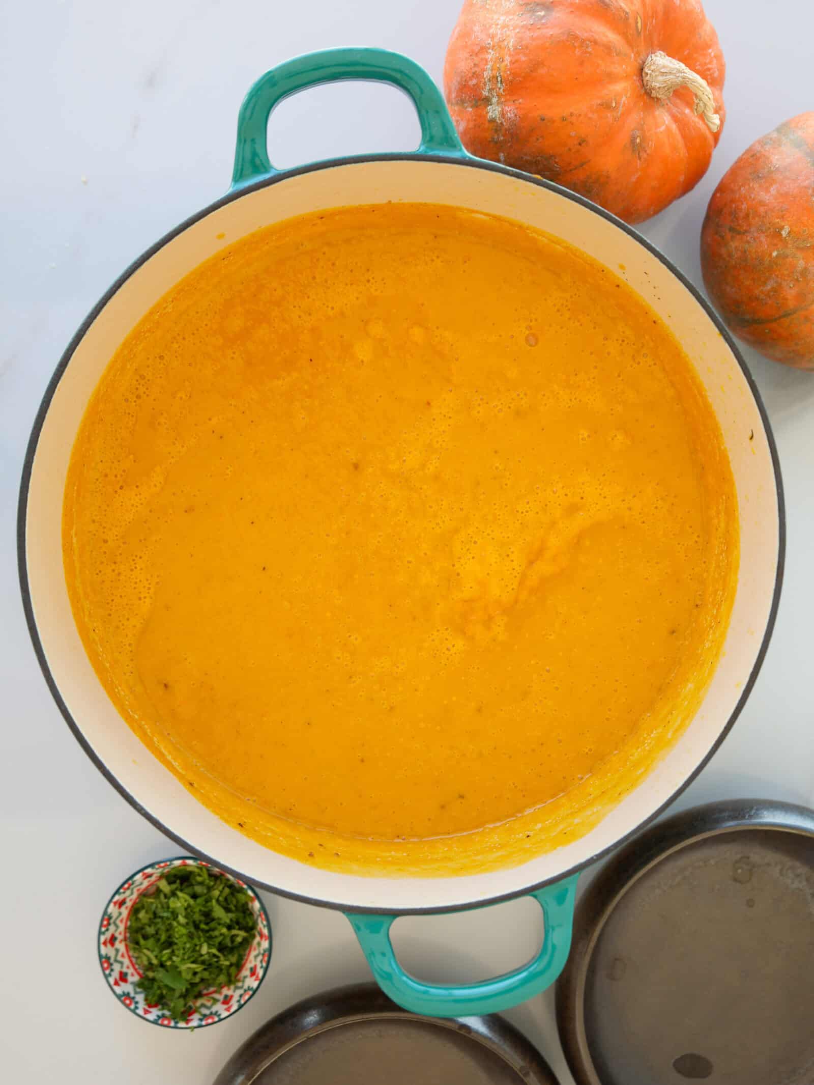 A pot of creamy pumpkin soup sits on a white surface, with two whole pumpkins nearby, a small bowl of chopped herbs, and a couple of round metal lids visible next to the pot.