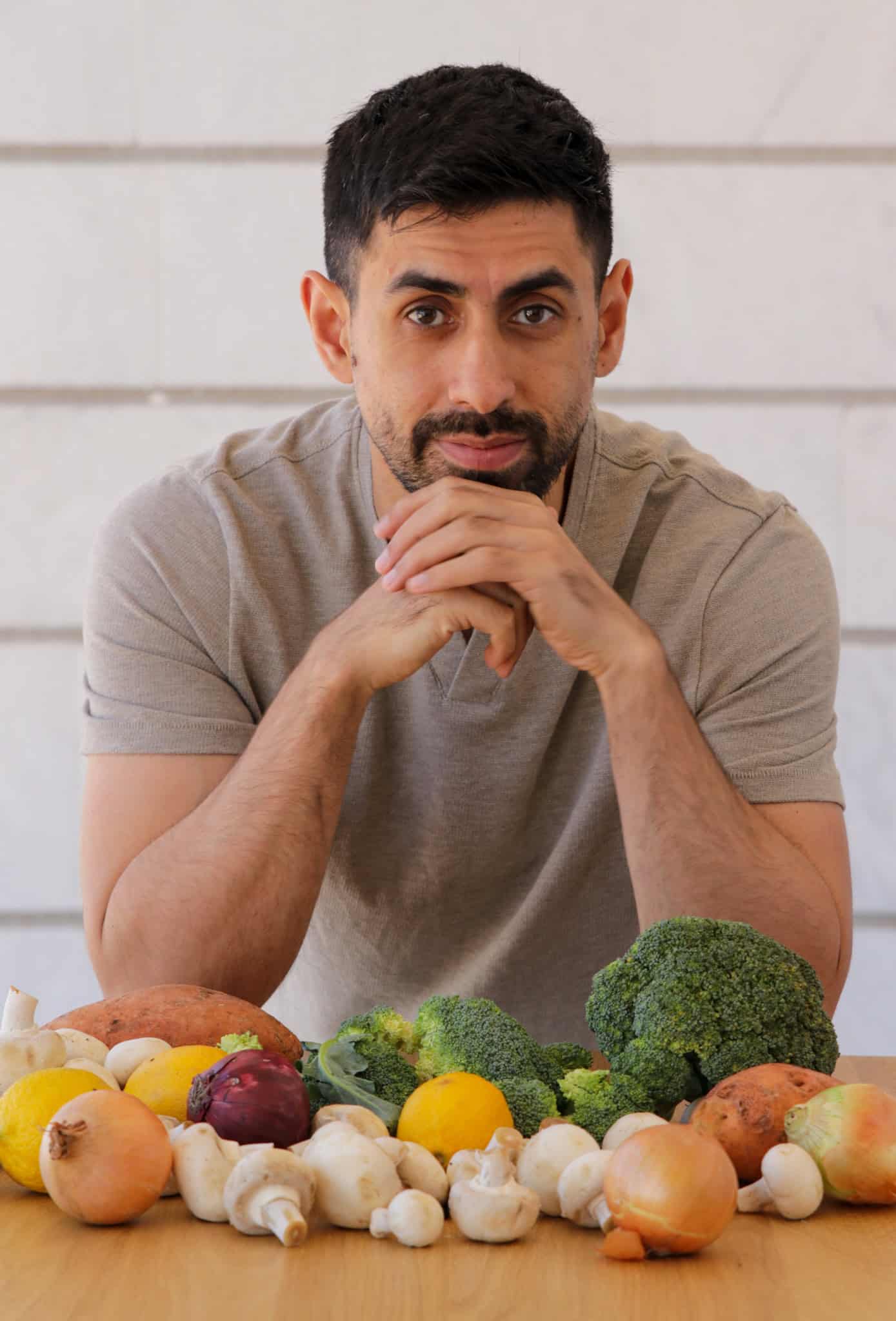 A man with dark hair and a beard sits at a table with his hands clasped, surrounded by various vegetables including broccoli, onions, mushrooms, lemons, and sweet potatoes.