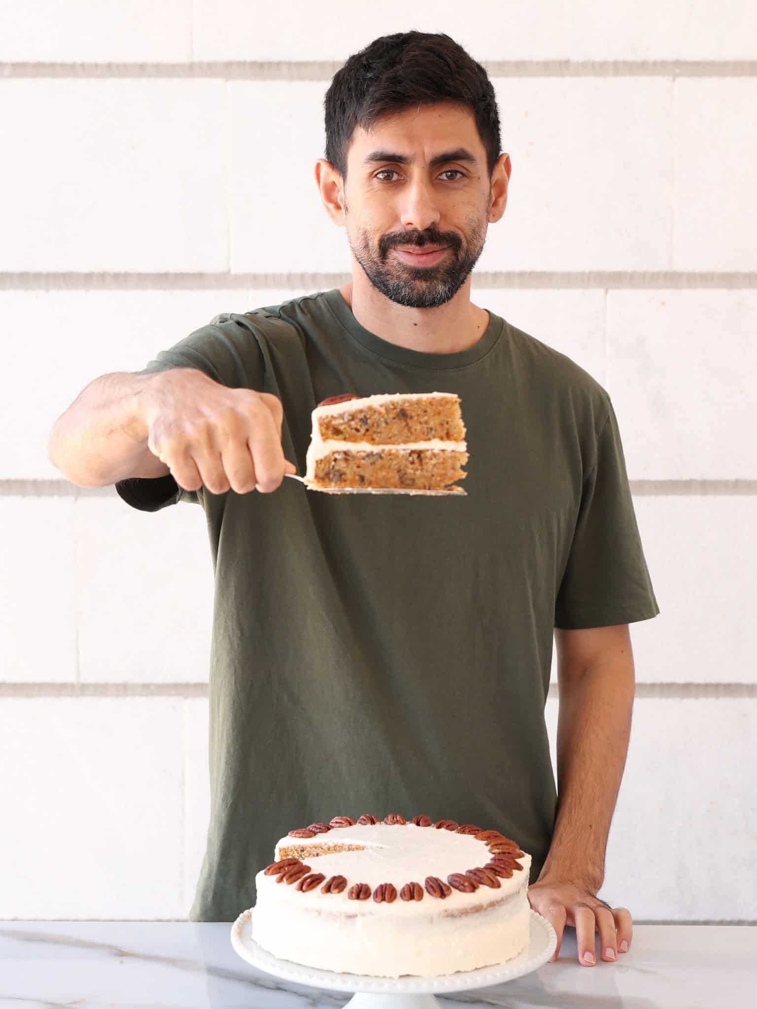 A man with dark hair and a beard, wearing a green t-shirt, holds a slice of cake on a spatula above a decorated round cake on a marble surface. The cake is topped with pecans and white frosting.