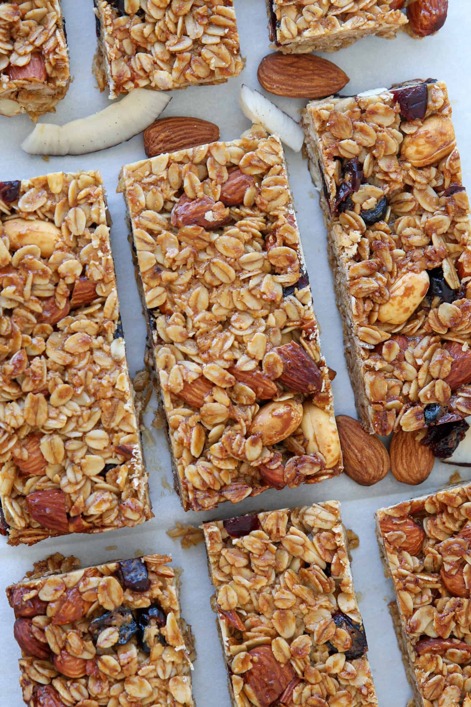 Close-up of healthy homemade granola bars on parchment paper, packed with oats, whole almonds, coconut flakes, and dried fruit pieces. The bars are rectangular and surrounded by loose nuts and coconut.