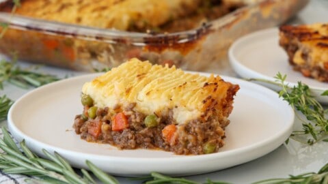 Best Shepherd’s Pie Recipe | Classic & Foolproof A slice of shepherd’s pie with a golden mashed potato topping sits on a white plate, showing layers of seasoned ground meat, peas, and carrots. Fresh rosemary sprigs are arranged around the plate.