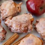 Glazed apple fritters with shiny red apples and cinnamon sticks on a white surface. The fritters are golden brown and covered in a sweet icing.