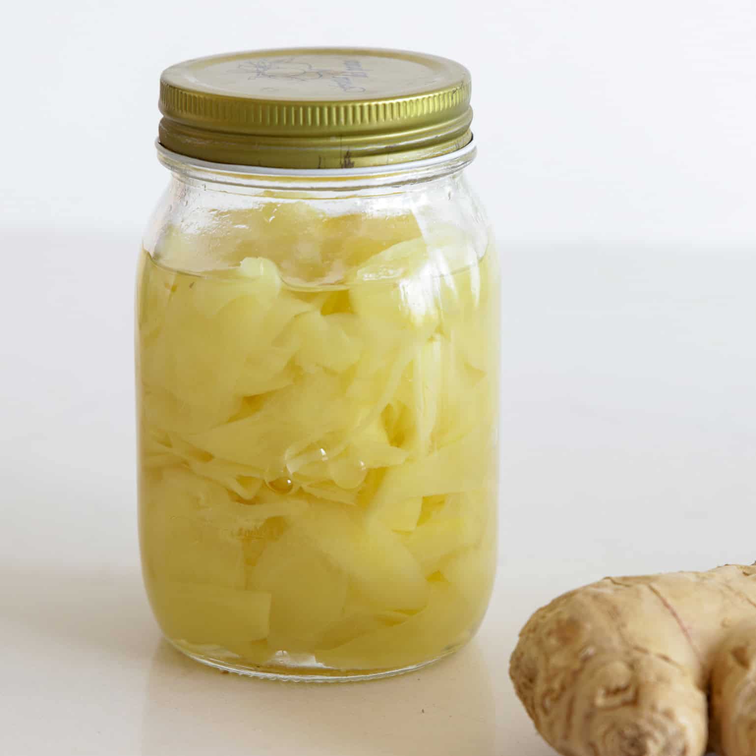 A glass jar with a gold lid filled with pickled ginger slices in liquid, sitting on a white surface. A piece of fresh ginger is partially visible in the bottom right corner.