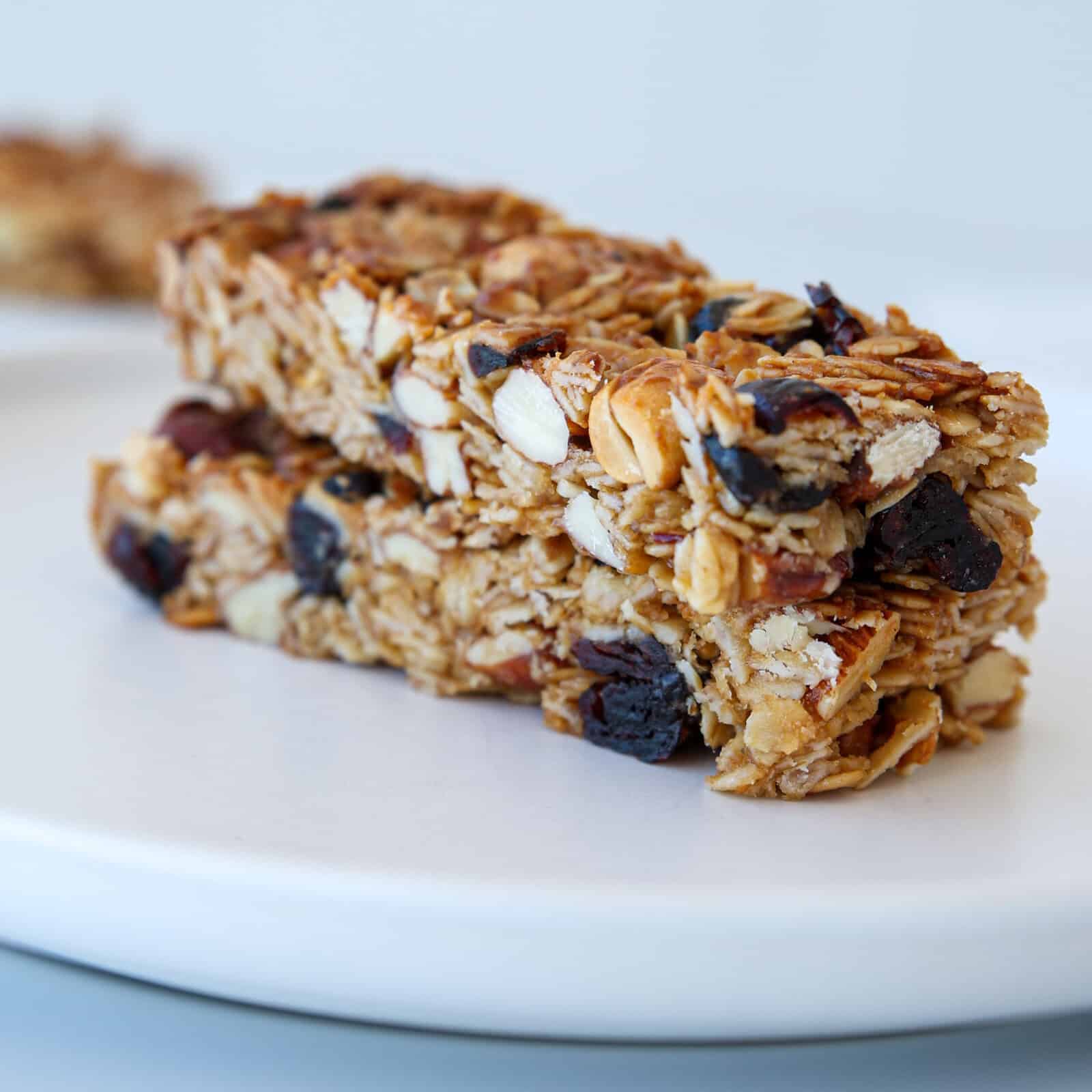 Two homemade granola bars with oats, nuts, and dried fruit are stacked on a white plate. The bars have a crunchy texture and are set against a plain, light background.