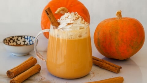 A glass mug of pumpkin spice latte topped with whipped cream and a cinnamon stick, surrounded by whole pumpkins, cinnamon sticks, and a small bowl on a white surface.