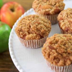 Four streusel-topped muffins on a white plate, with green and red apples in the background on a wooden surface.