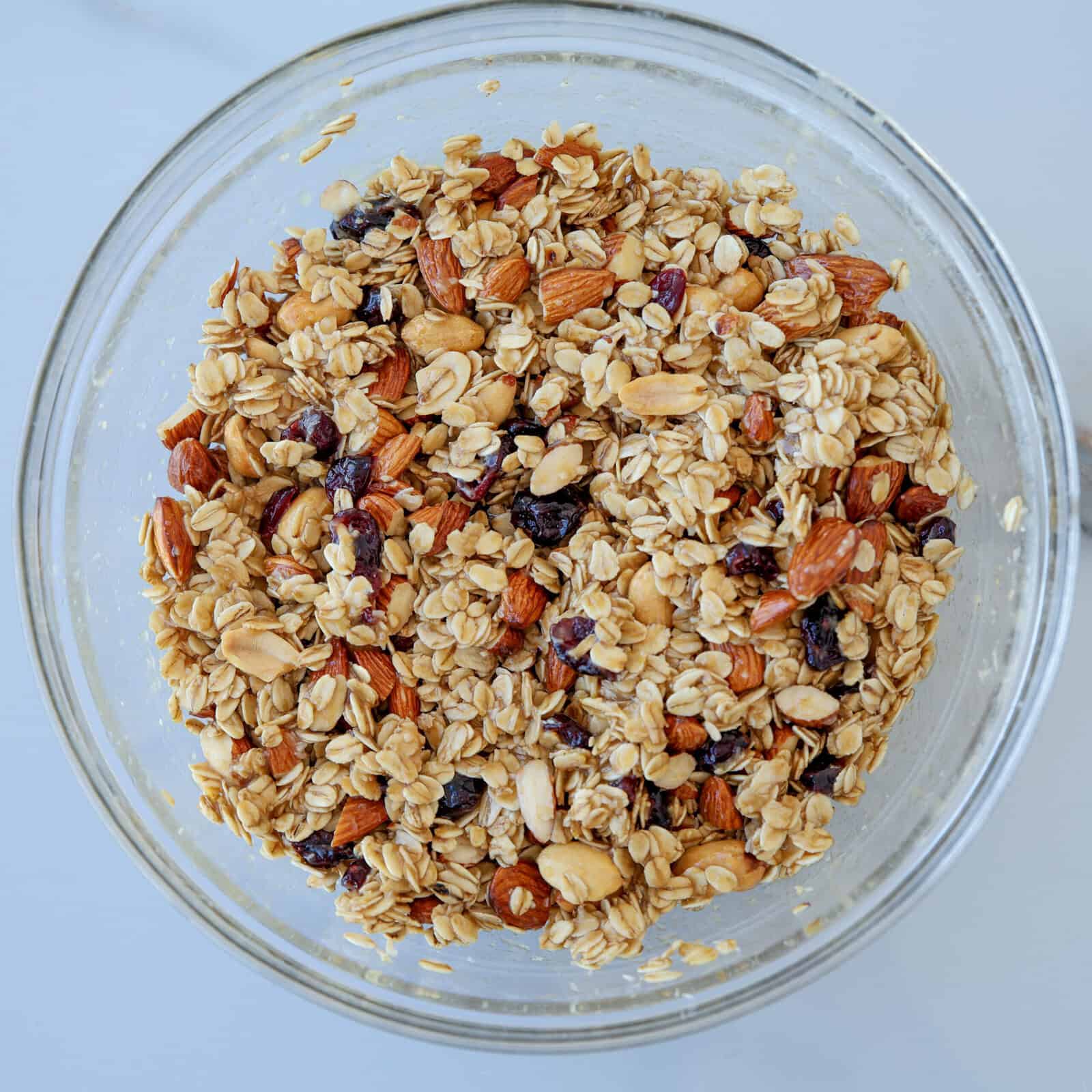A glass bowl filled with homemade granola containing rolled oats, almonds, peanuts, and dried berries, set on a light-colored surface.
