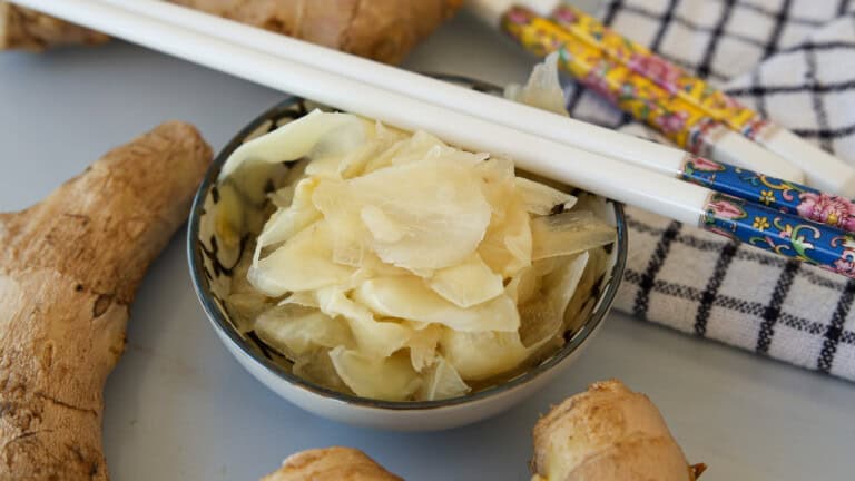 A small bowl of pickled ginger slices is surrounded by fresh ginger roots, white chopsticks with colorful patterns, and a checkered cloth on a light surface.