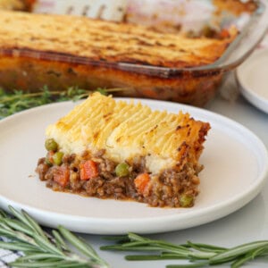 A slice of shepherd’s pie with a layer of mashed potatoes on top, browned and ridged, sits on a white plate. The filling beneath includes ground meat, peas, and carrots. A baking dish and rosemary sprigs are in the background.