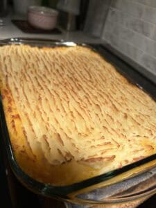 A glass baking dish filled with golden-brown shepherd’s pie, featuring a textured, ridged layer of mashed potatoes on top, sits on a kitchen counter under warm lighting.